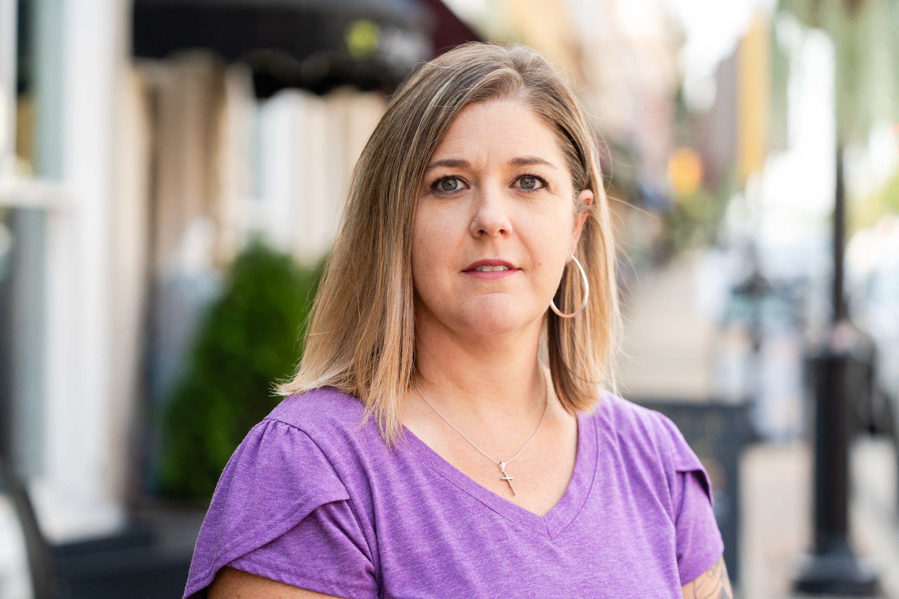 A woman in purple shirt standing on street next to trees.