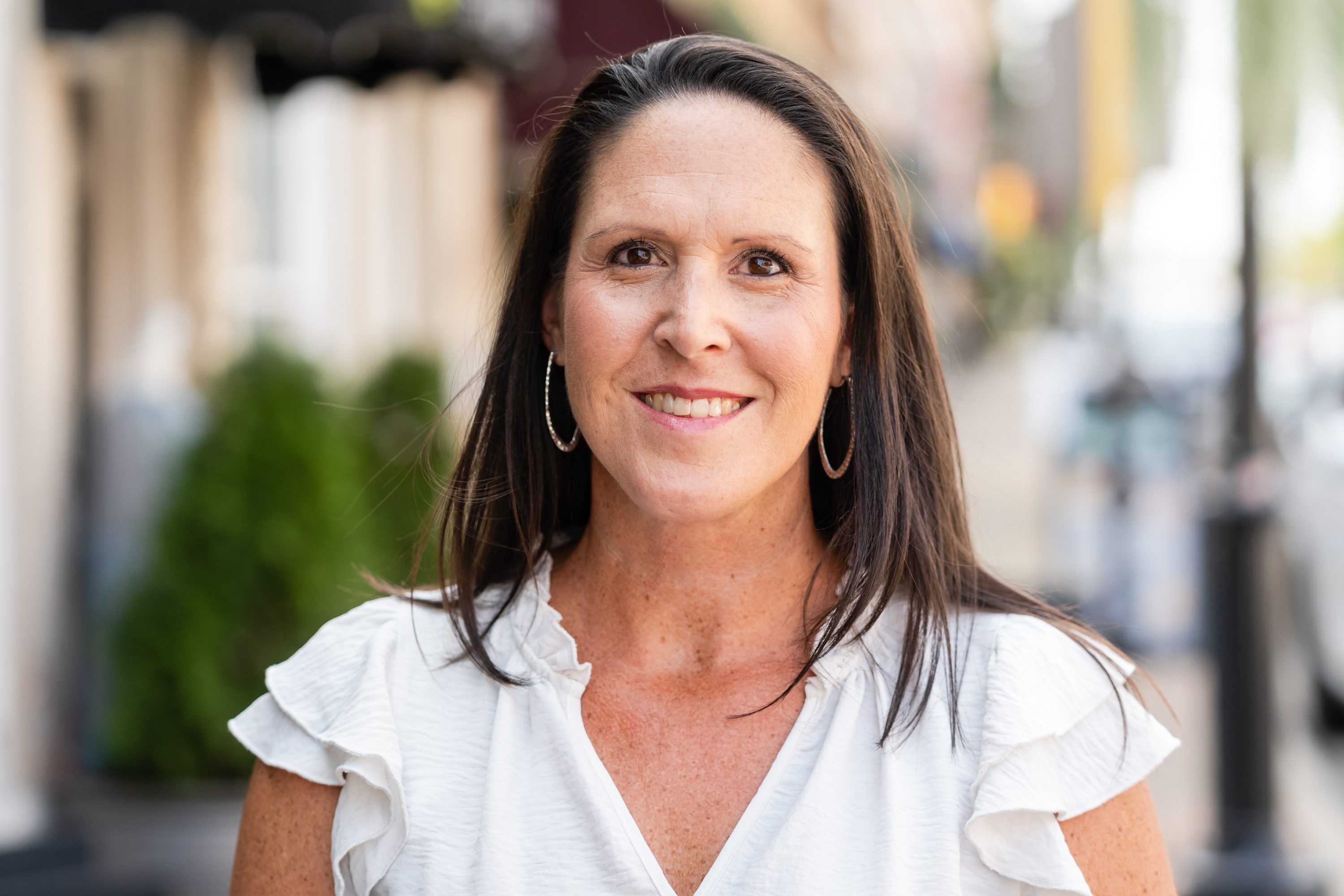 A woman in white shirt standing on street next to trees.